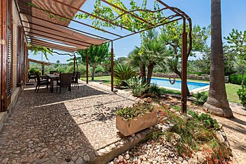 A sunny outdoor patio with a shaded dining area, cobblestone floor, lush garden, palm trees, and a sparkling blue swimming pool in the background.