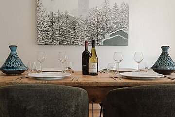 A set wooden dining table featuring wine bottles, plates, glasses, blue ceramic tagines, and a winter painting on the wall behind.