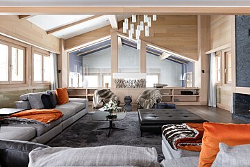 A luxurious chalet living room with vaulted wood ceilings, modern gray sofas, orange accents, and a contemporary chandelier creating a cozy, stylish space.