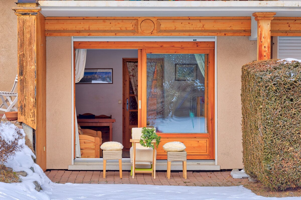 A house with wooden trim and a glass sliding door opens to a snowy patio with stools and a plant, flanked by a hedge.