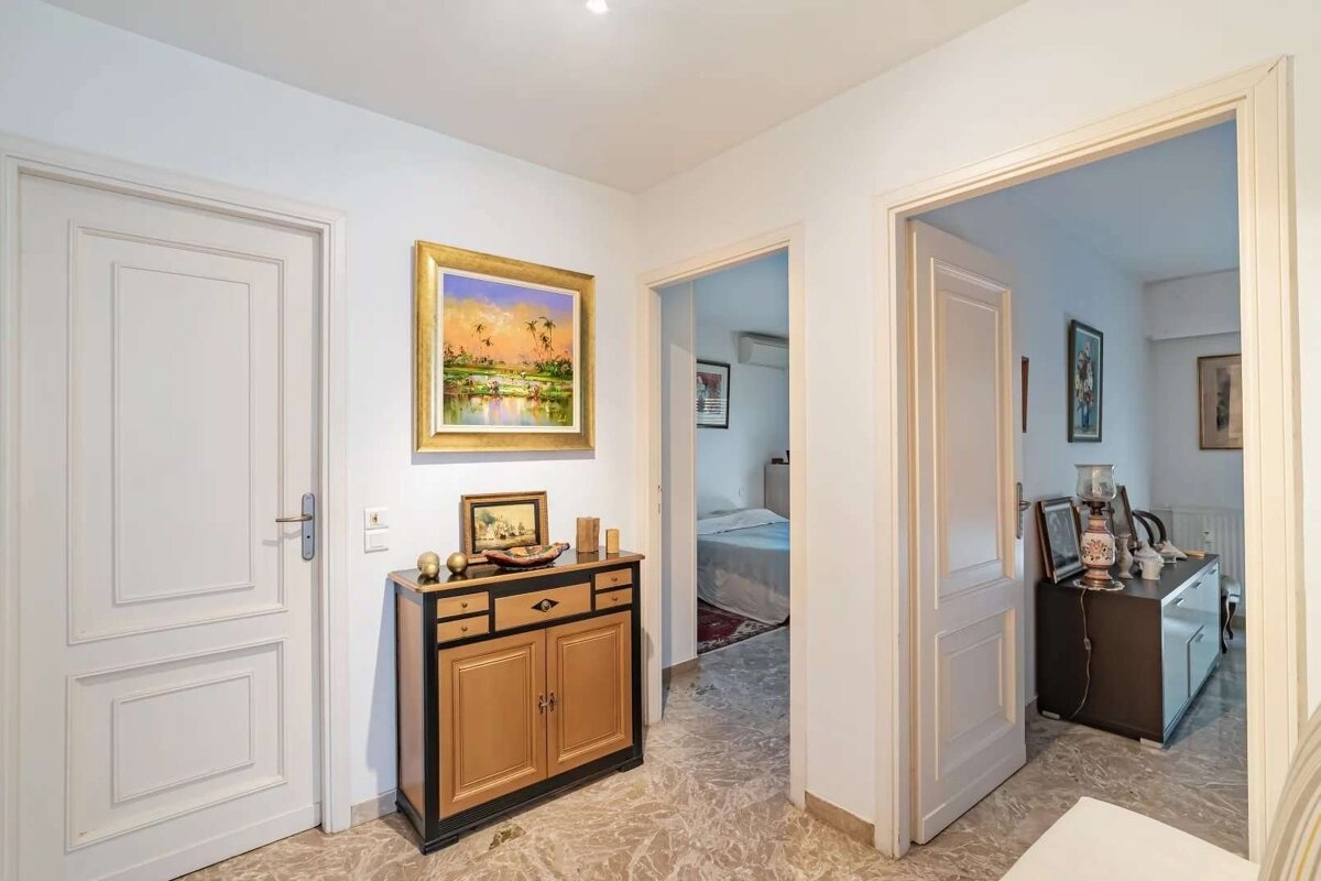 A bright hallway features a large painting, a wooden cabinet, and marble floors. Two open doorways reveal glimpses into adjacent rooms.