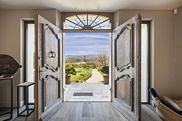 Open wooden double doors look out onto a scenic, sunlit path through a lush green garden towards distant hills.