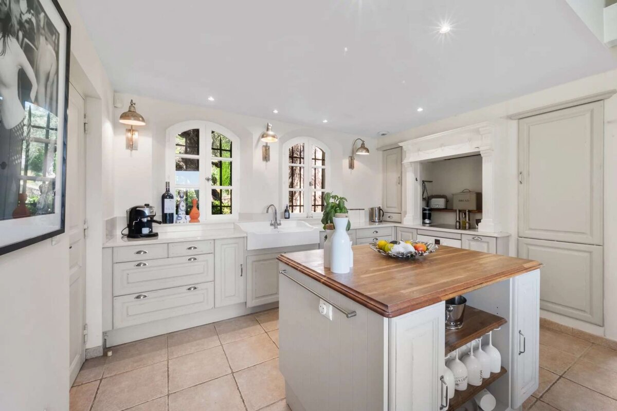 A bright, classic white kitchen with arched windows, a farmhouse sink, a central wooden-topped island, and tiled floors.