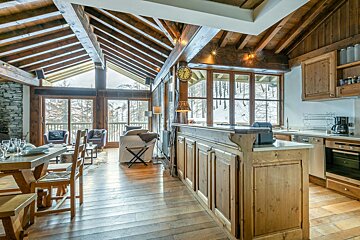 Spacious wooden chalet interior with open-plan living, dining, and kitchen areas, featuring large windows offering a snowy mountain view.