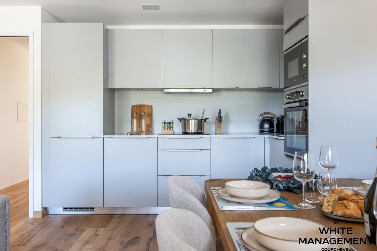 A sleek, modern kitchen with light gray cabinetry and integrated appliances, opening to a dining area with a wooden table set for guests.