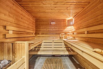 A warm, inviting wooden sauna with multi-level benches, a bucket, and a thermometer on the wall. Sunlight streams through a vent.