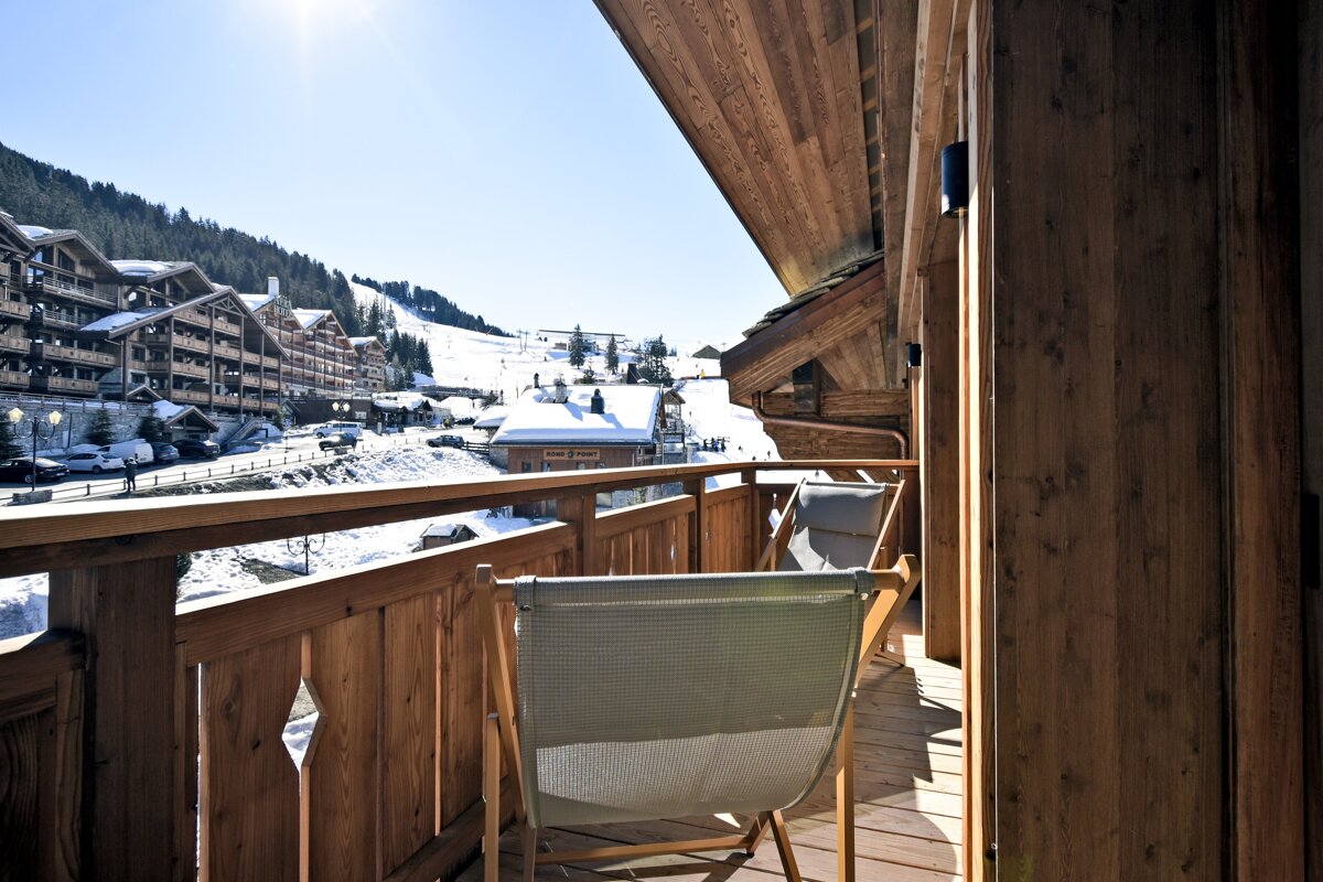 View from a wooden balcony with deck chairs overlooking a snowy ski resort and alpine chalets on a sunny day.