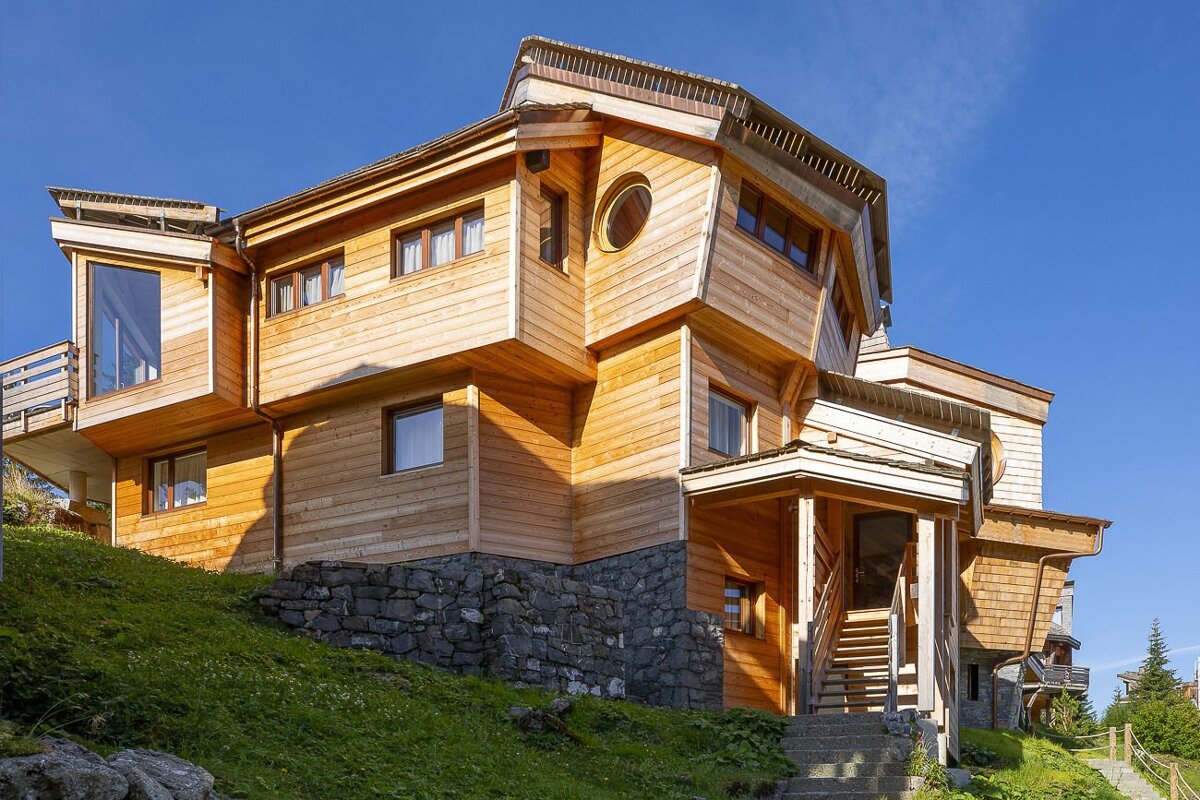A distinct, multi-faceted wooden house with varied angles, a round window, and stone foundation, set on a green hillside under a clear blue sky.