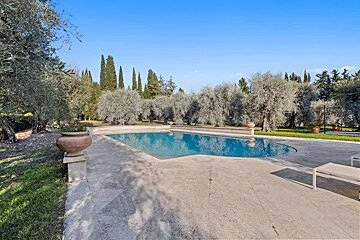 A serene outdoor swimming pool with a paved patio, surrounded by mature olive and cypress trees under a clear blue sky on a sunny day.
