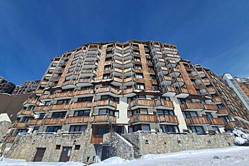 A massive, tiered ski resort building, featuring wood shingles, balconies, and a stone base, standing tall against a bright blue sky in the snow.