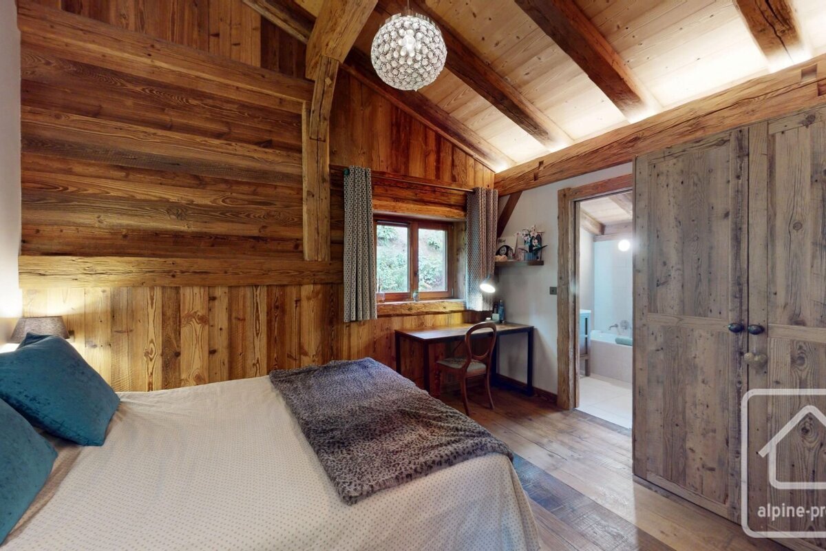A rustic, wood-paneled bedroom features a bed with blue pillows, a desk by a window, and a sparkling chandelier. A bathroom is visible beyond an open doorway.