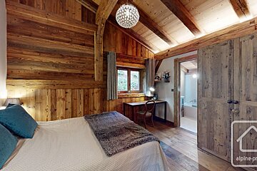 A rustic, wood-paneled bedroom features a bed with blue pillows, a desk by a window, and a sparkling chandelier. A bathroom is visible beyond an open doorway.