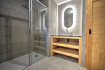 Modern bathroom with a glass-enclosed shower, stone tiles, a wooden vanity, and a backlit oval mirror.