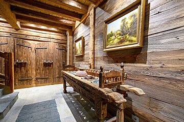 A rustic room with exposed wooden beams, plank walls, and a carved wooden console table with pottery. Landscape paintings hang above a stone floor.