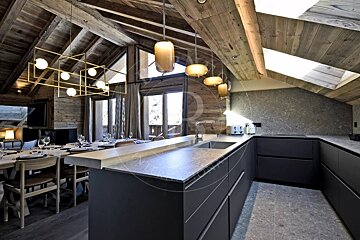 Modern chalet kitchen and dining area featuring dark cabinetry, stone countertops, wooden ceilings, and contemporary lighting.