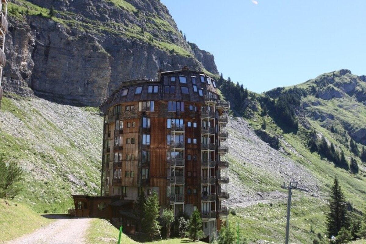 A large, multi-story wooden chalet with many balconies and snow-covered roofs is nestled in a snowy mountain landscape under a blue sky.