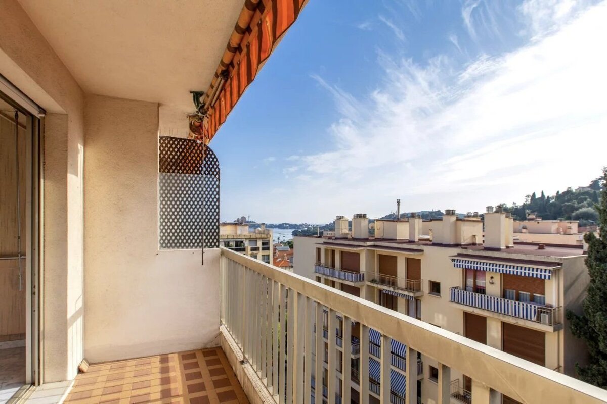 Sunny apartment balcony with tiled floor and orange awning, overlooking city buildings and a distant sea under a bright blue sky.