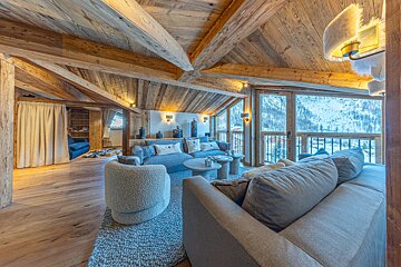 Modern alpine chalet interior with rustic wooden beams, plush seating, and large windows showcasing snowy mountain views.