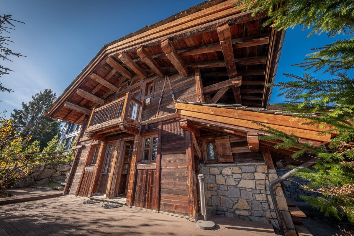 Rustic wooden chalet with a stone foundation, featuring a small balcony and wooden deck, surrounded by trees under a bright blue sky.