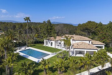 Aerial view of a luxurious white house with a pool, surrounded by palm trees, green lawns, and forests under a clear blue sky.