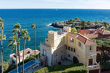 A grand villa with red roofs and palm trees on a cliff, overlooking a bright blue ocean with a rocky coastline under a clear sky.