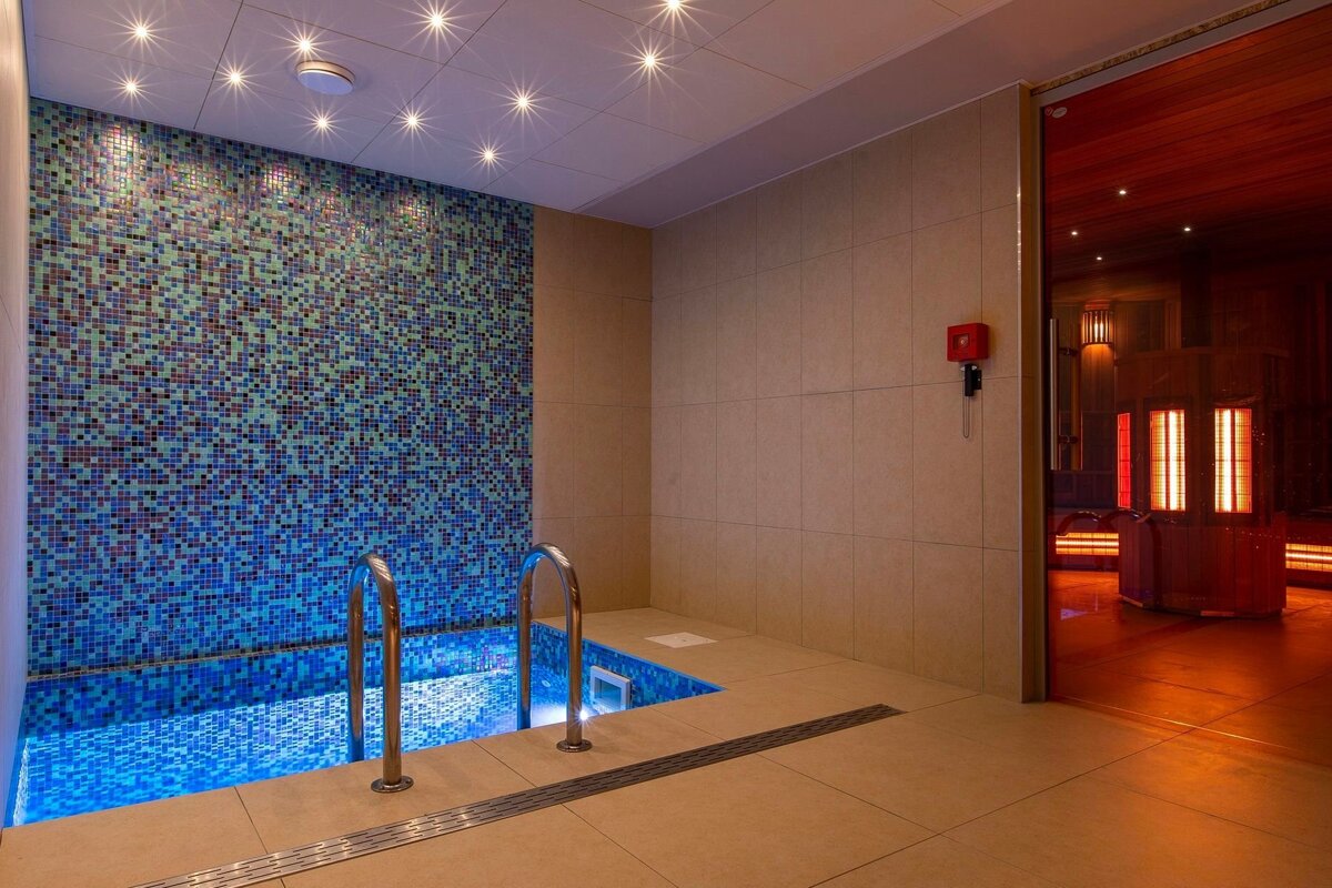 A modern spa room with a blue mosaic plunge pool, bright ceiling lights, and a warm, red-lit sauna visible through an open doorway.