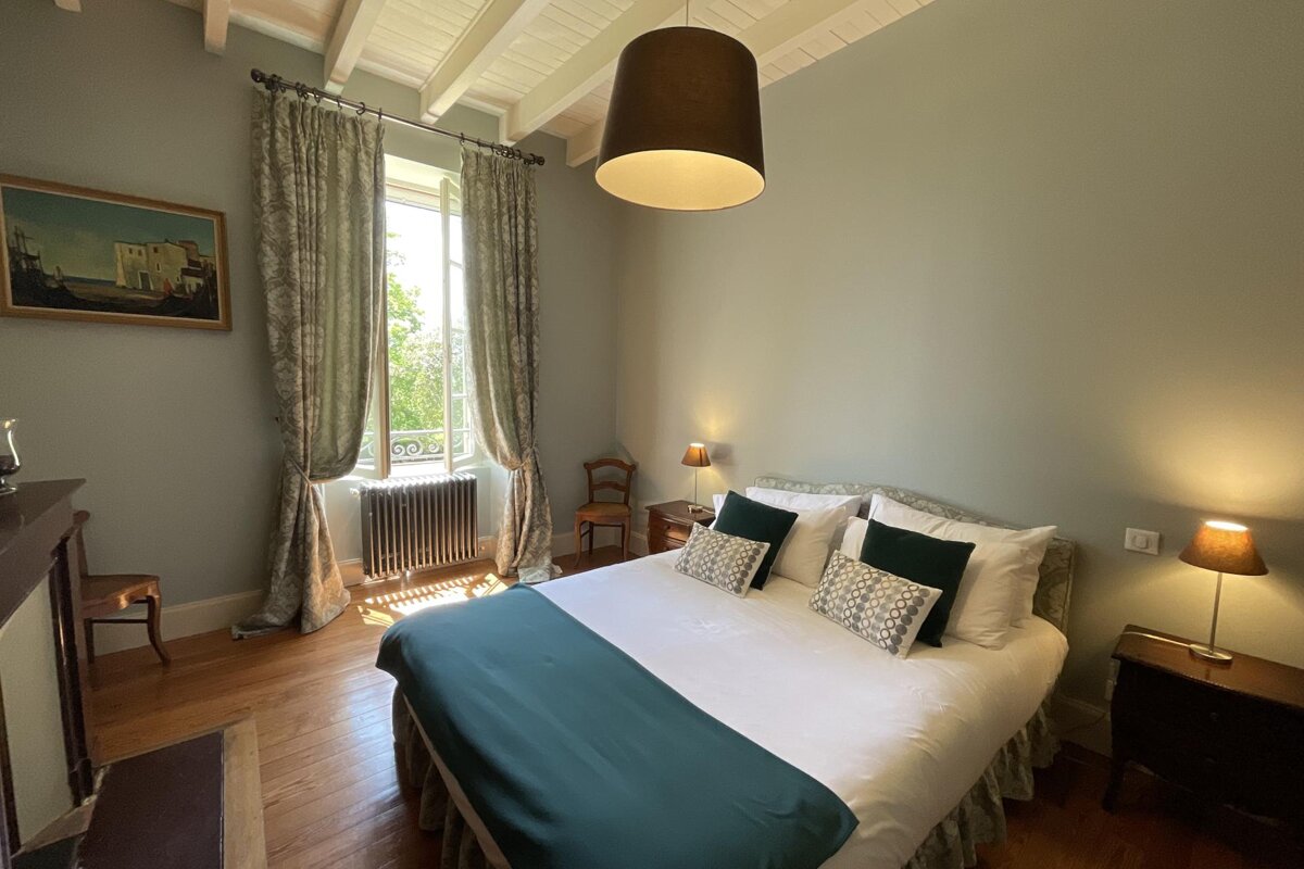A bright, inviting bedroom featuring a large bed with green and white bedding, light green walls, hardwood floors, and natural light from an open window.