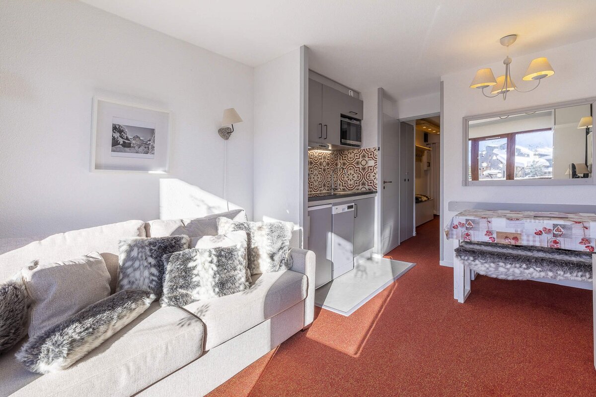 Sunny studio apt with beige sofa & furry pillows, gray kitchenette, patterned backsplash, dining table, red carpet. Mountain views reflected.