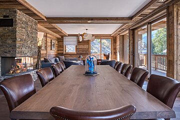 A cozy, rustic chalet dining room with a long wooden table, leather chairs, stone fireplace, and a living area, offering mountain views.