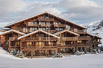 A grand, multi-story wooden chalet blanketed in snow, nestled amidst snowy mountains under a cloudy sky.