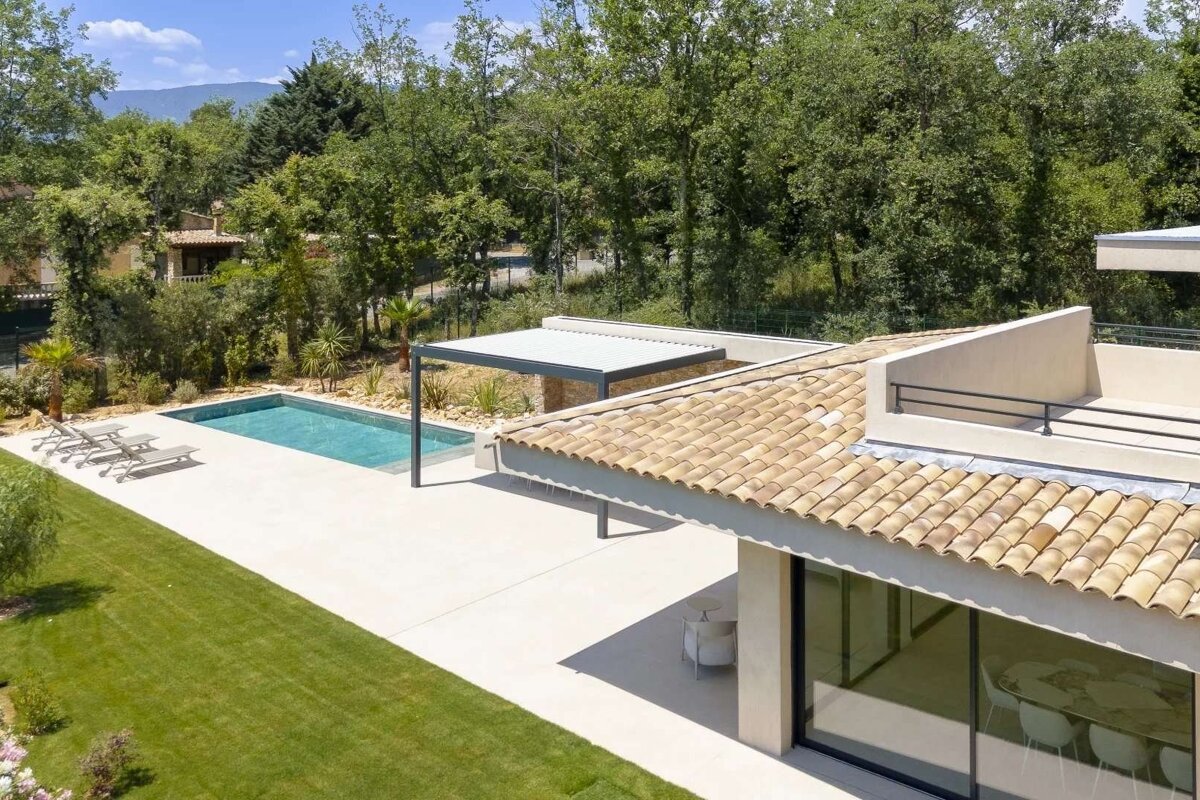 Aerial view of a modern villa featuring a tiled roof, sparkling swimming pool, concrete patio, and a vibrant green lawn amidst a wooded landscape.