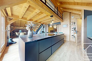 Spacious modern chalet interior with a black kitchen island, exposed wooden beams, and extensive light wood finishes. A dining area, mezzanine, and wine rack are visible.