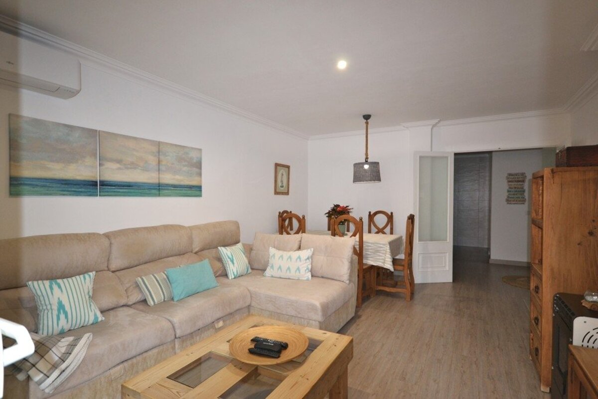 A bright living/dining room with a large L-shaped sofa, wooden coffee table, dining set, ocean artwork, and light wood floors.