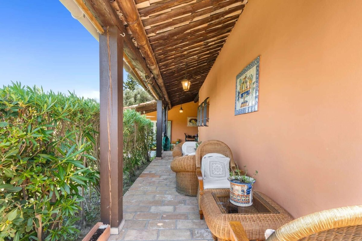 Covered patio with wicker chairs, orange wall, wooden beams, paved floor, and green hedge. Blue sky above.