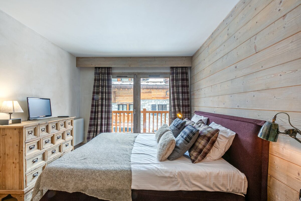 Cozy bedroom featuring a wooden accent wall, a bed with plaid pillows, a TV on a rustic dresser, and a balcony door.