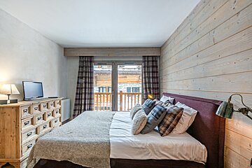 Cozy bedroom featuring a wooden accent wall, a bed with plaid pillows, a TV on a rustic dresser, and a balcony door.