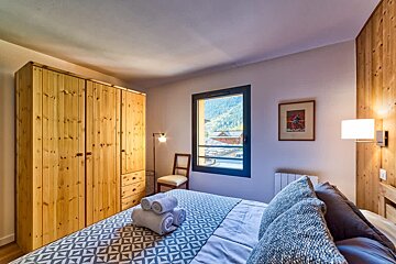 Cozy alpine bedroom with a large wooden wardrobe, patterned blue bedding, and a window offering mountain views.
