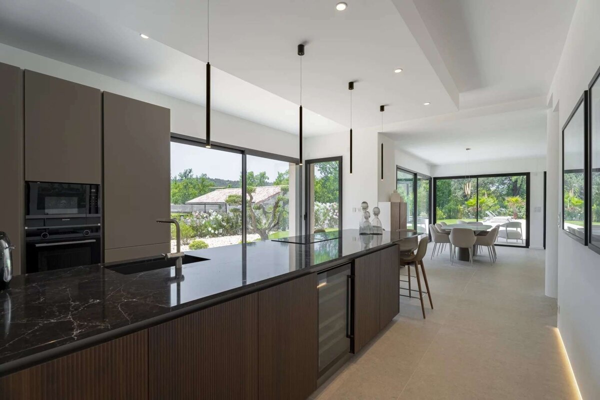 A modern, open-plan kitchen with dark cabinetry, a black marble island, and large windows overlooks a green outdoor space. A dining area is visible beyond.
