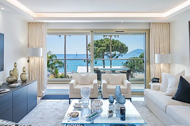 A modern, bright living room with white furnishings, stylish decor, and a stunning ocean view from a large balcony with palm trees.