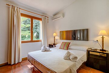 A bright, inviting bedroom featuring a double bed, white linens, terracotta floor, window with greenery view, bedside lamps, and colorful wall art.