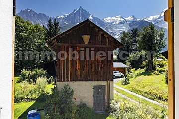A rustic wooden building stands before majestic snow-capped mountains and green trees under a clear blue sky, framed by a window.