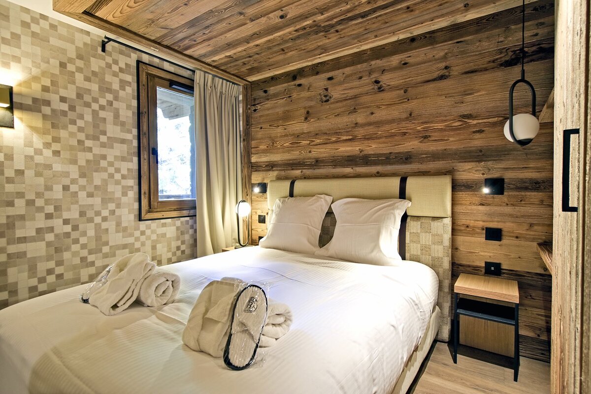 Cozy modern bedroom with rustic wood paneling, white bedding, bathrobes, and slippers. A window provides natural light, creating a warm ambiance.