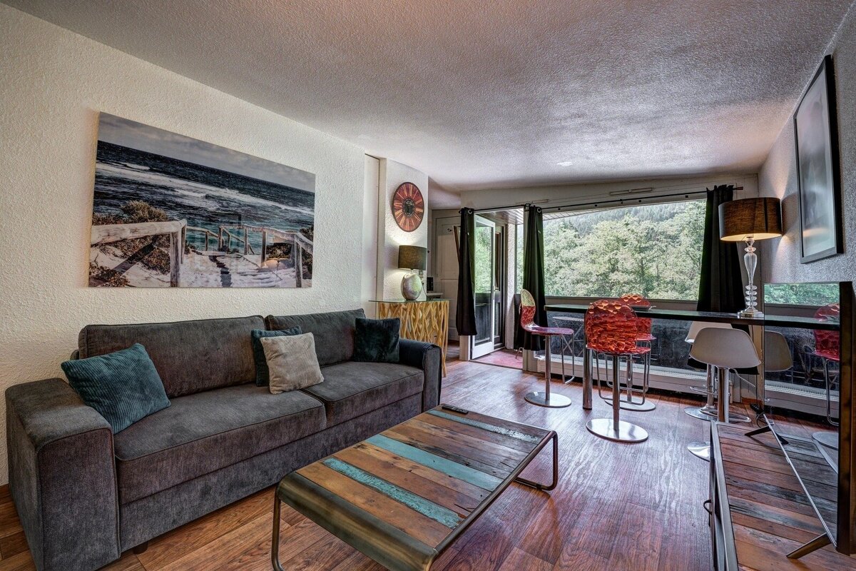 Modern living room with a grey sofa, rustic coffee table, large beach wall art, and a dining bar looking out to forest views.