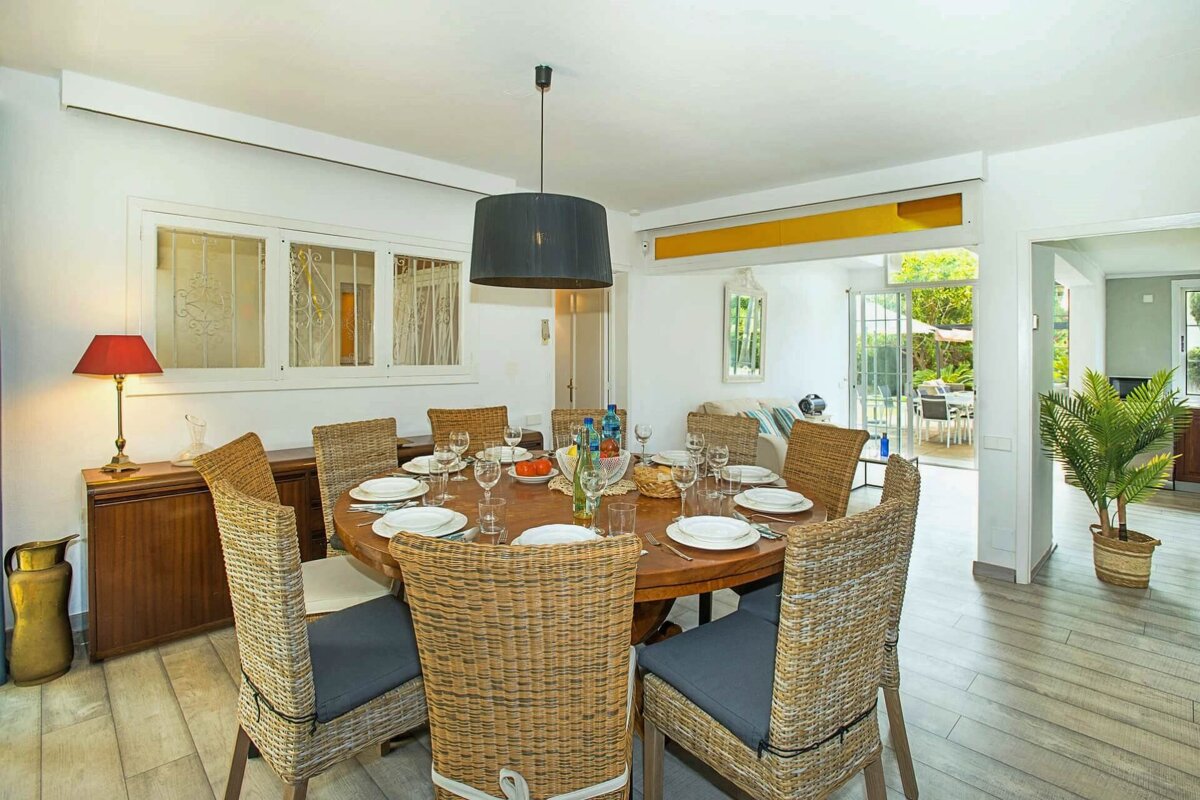 A bright dining room features a large, set round wooden table with wicker chairs, illuminated by a modern pendant light. It opens to a patio view.