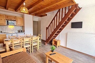 Rustic wooden cabin interior featuring a dining table, open staircase, beamed ceiling, kitchen, and a cozy living area.