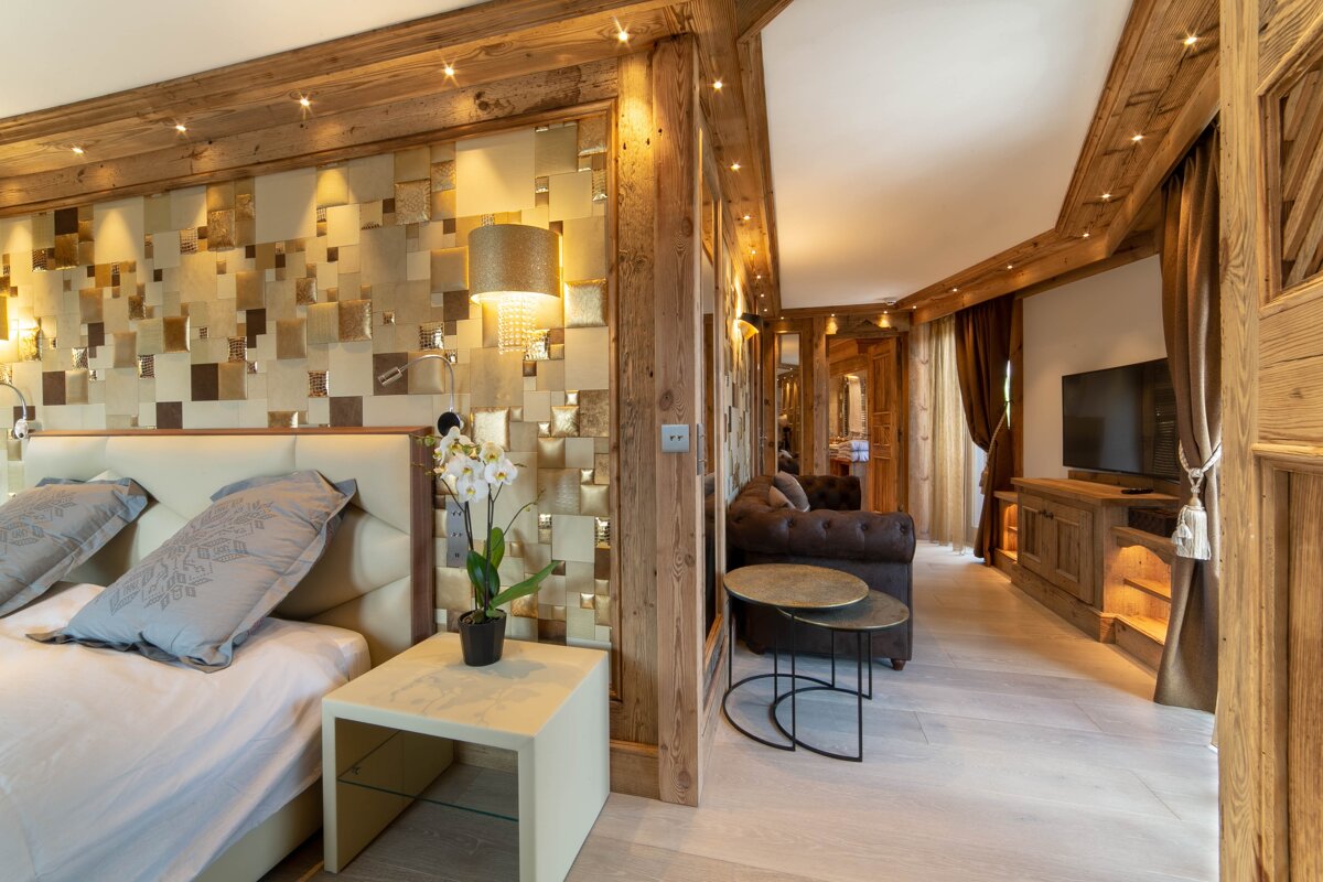 Luxury chalet bedroom and living space. Wooden walls frame a bed with a textured headboard wall, and a cozy lounge with a sofa and TV.