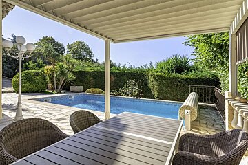 A sunny patio with a table and wicker chairs overlooks a sparkling blue swimming pool surrounded by lush green hedges and trees.