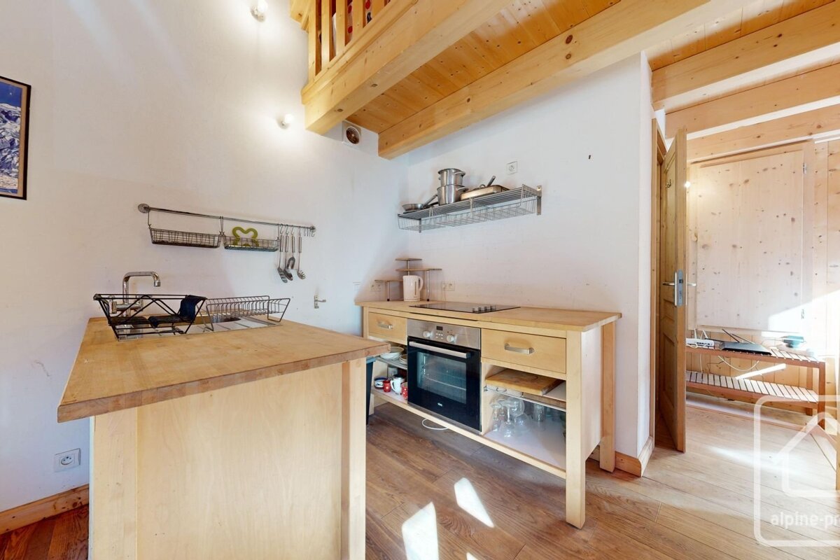 A bright, modern kitchen features light wood counters, cabinets, and exposed ceiling beams leading to a mezzanine. White walls, oven, and sink visible.