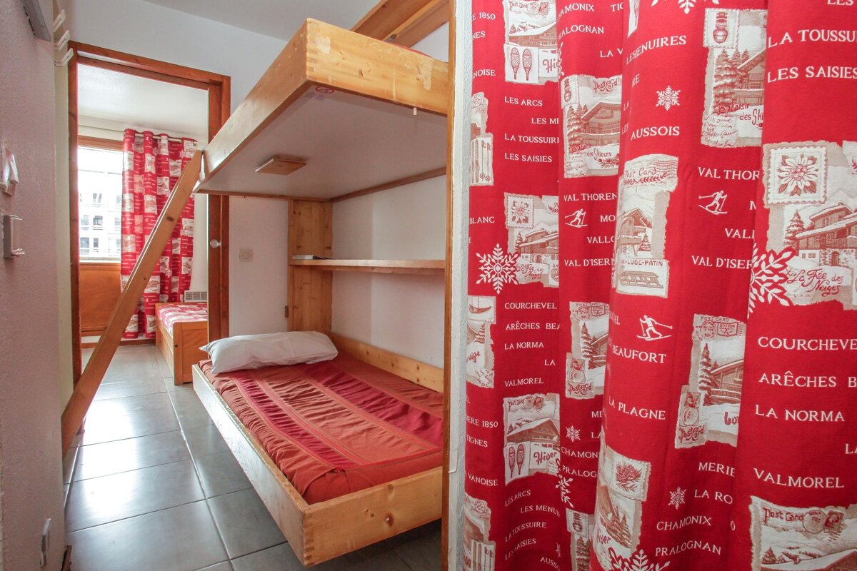 A cozy ski chalet bedroom featuring wooden bunk beds with red mattresses and a vibrant red curtain adorned with ski resort names and snowflakes.