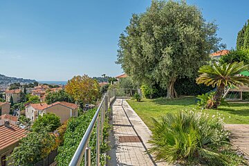 Sunny garden path overlooking a town with red roofs and the blue sea in the distance. Lush greenery and an olive tree on a bright day.